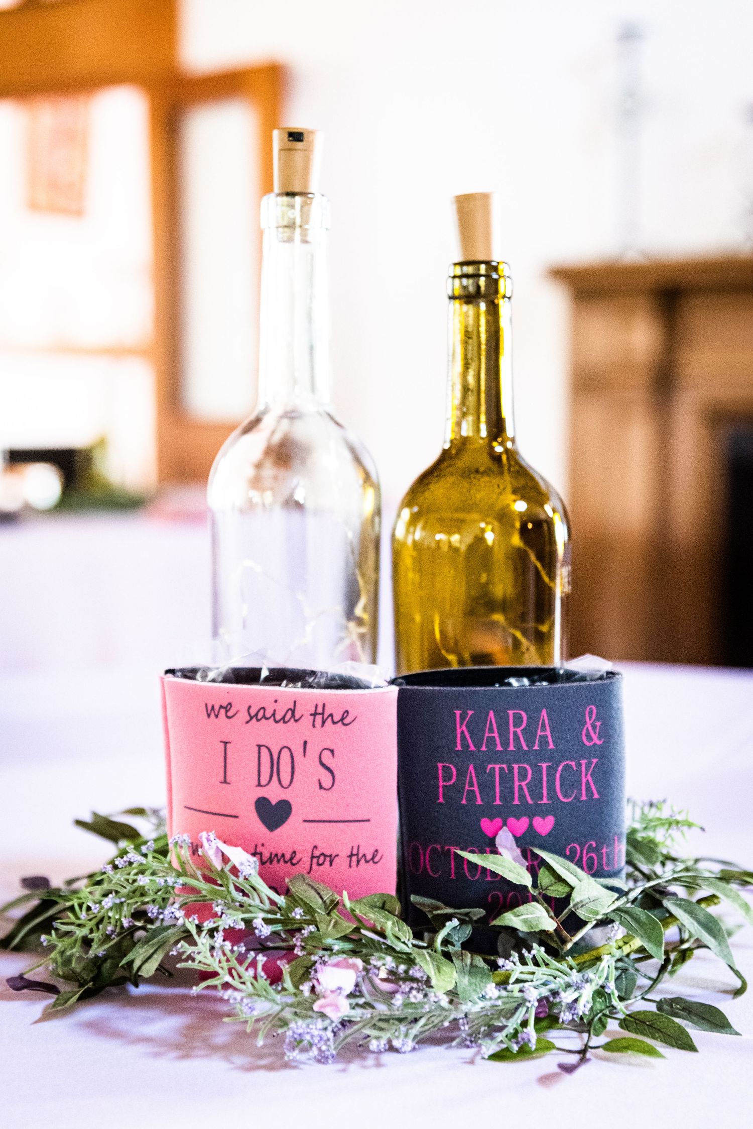Wedding wine bottles decorated with custom pink and black koozies surrounded by greenery.