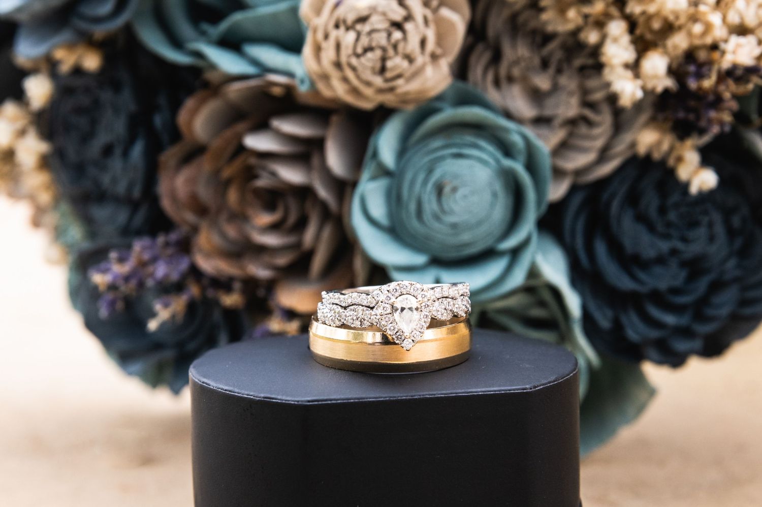 Wedding rings displayed on black stand with blue and neutral toned fabric flowers in background.