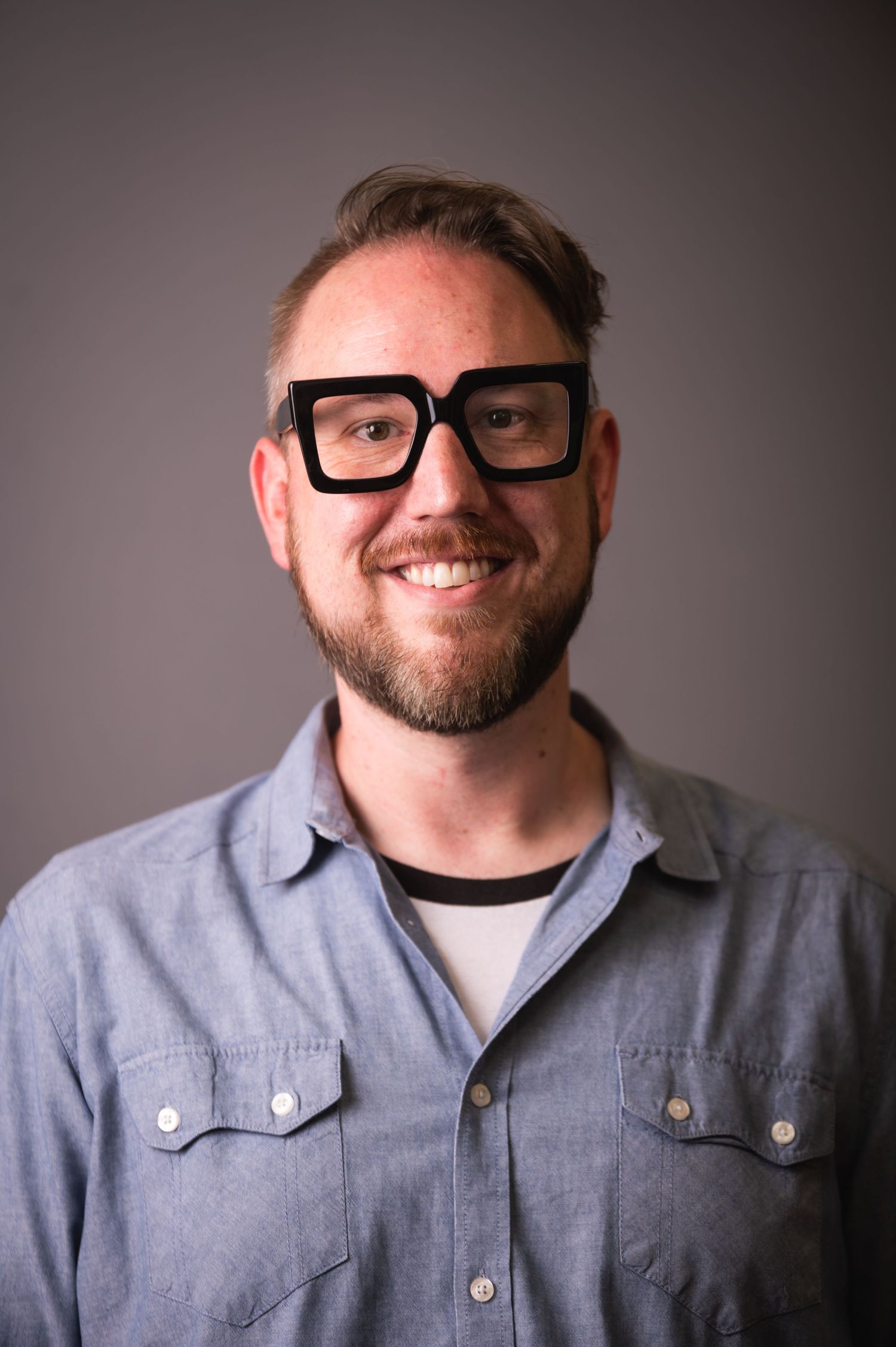 Portrait of person in gray button-up shirt and black-rimmed glasses smiling at camera.