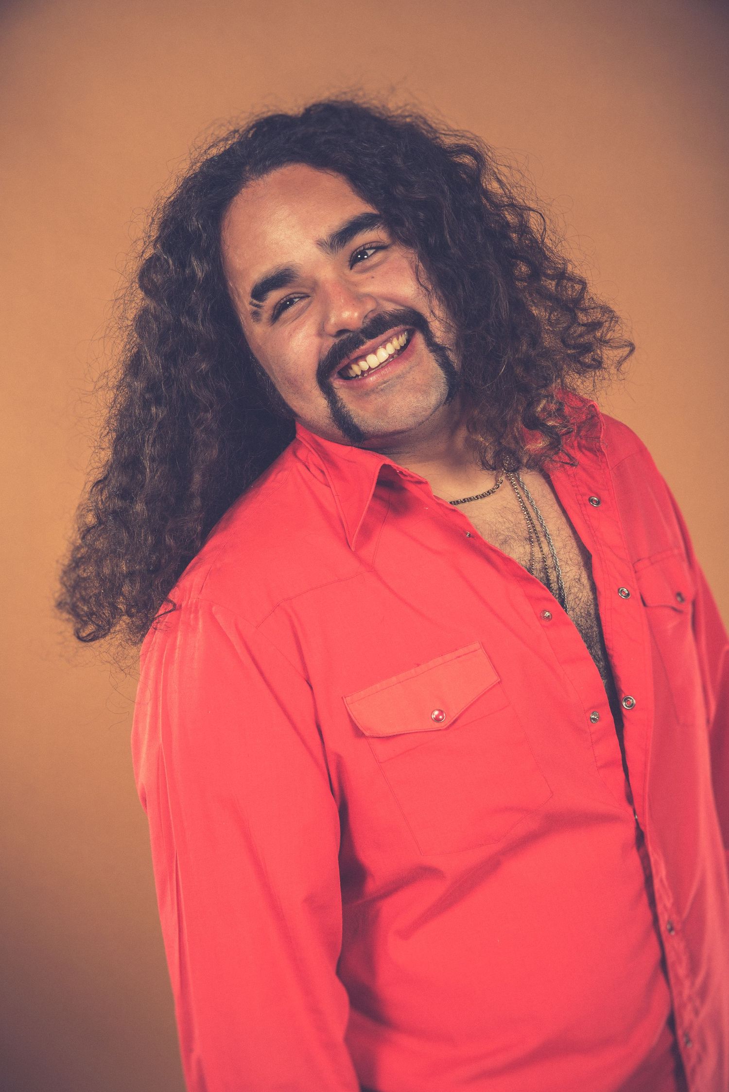 Portrait of someone with long curly hair and a mustache wearing a bright coral shirt against an orange backdrop.