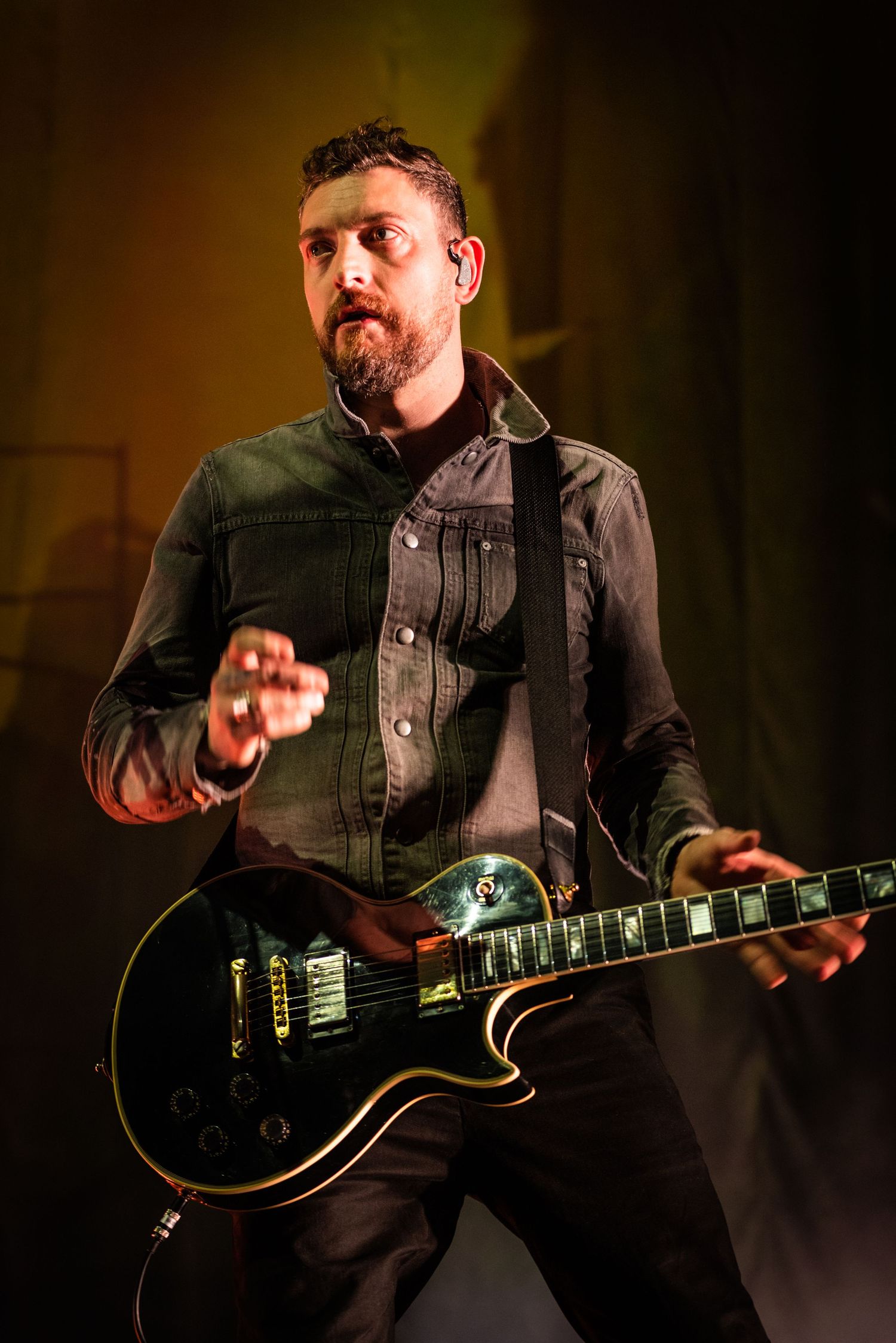 Musician performs on stage with a black electric guitar under dramatic amber lighting at a concert.