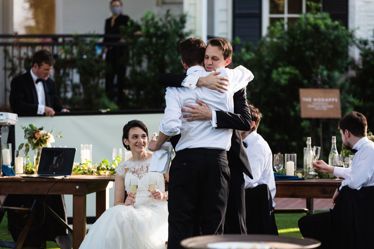 Emotional embrace during wedding reception with guests seated at outdoor tables.