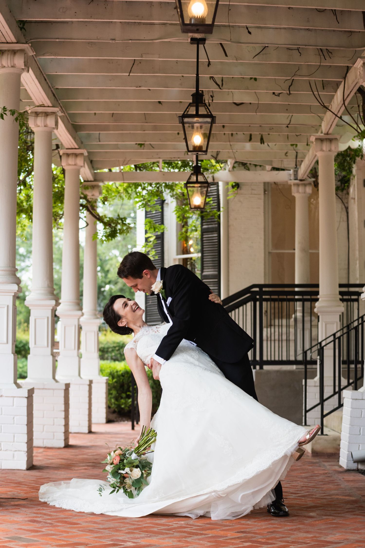 A dramatic wedding dip pose under a covered porch with hanging lanterns and classic columns.