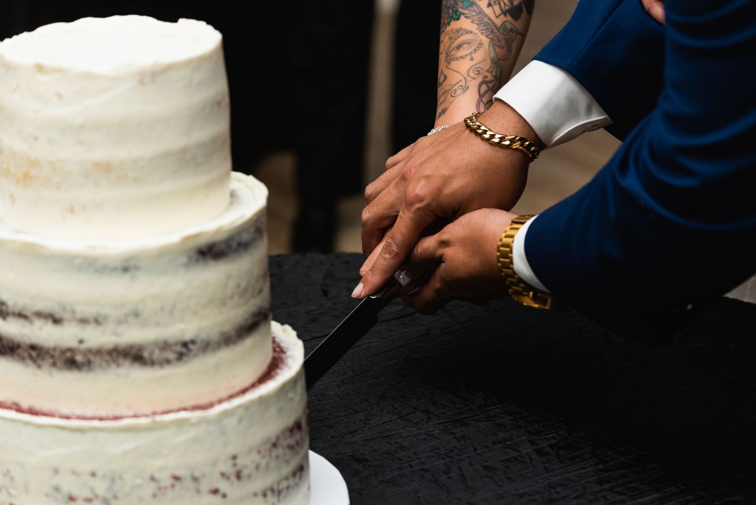 Close-up of hands cutting a three-tiered white wedding cake on black tablecloth.