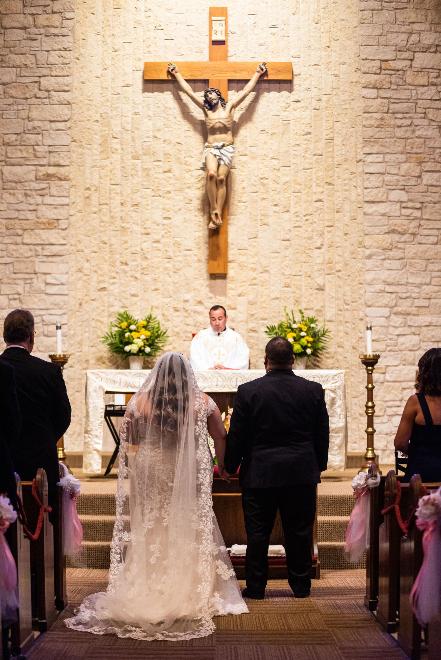 A wedding ceremony taking place in a Catholic church with a large crucifix and yellow floral arrangements.