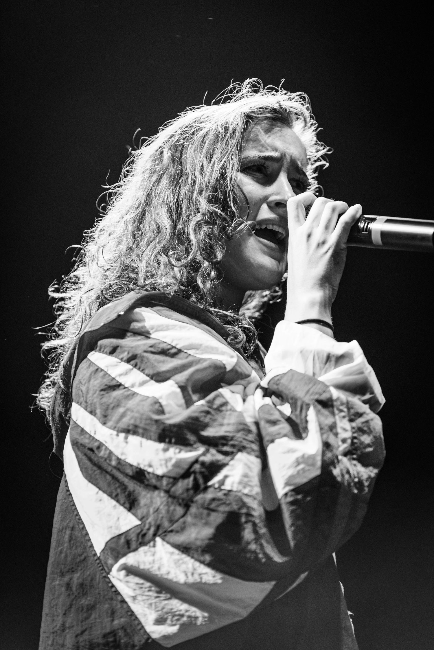 Concert performer with curly hair sings passionately into microphone wearing striped jacket in black and white photo.