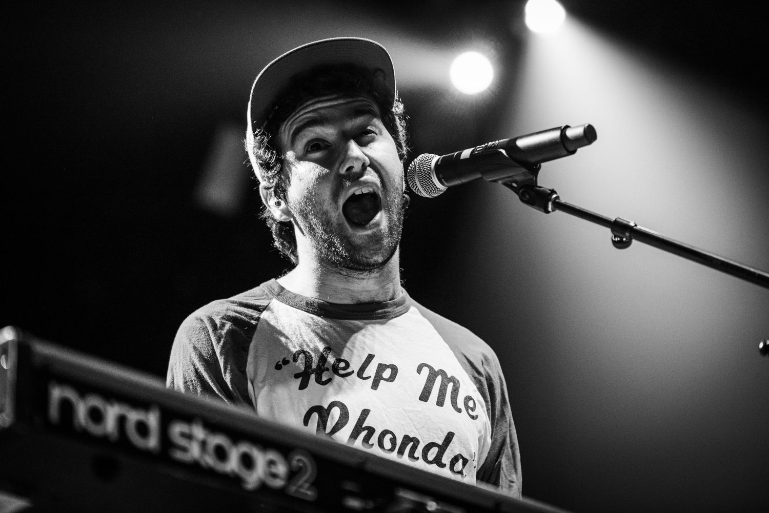 Black and white photo of a musician singing at a keyboard during a live performance.