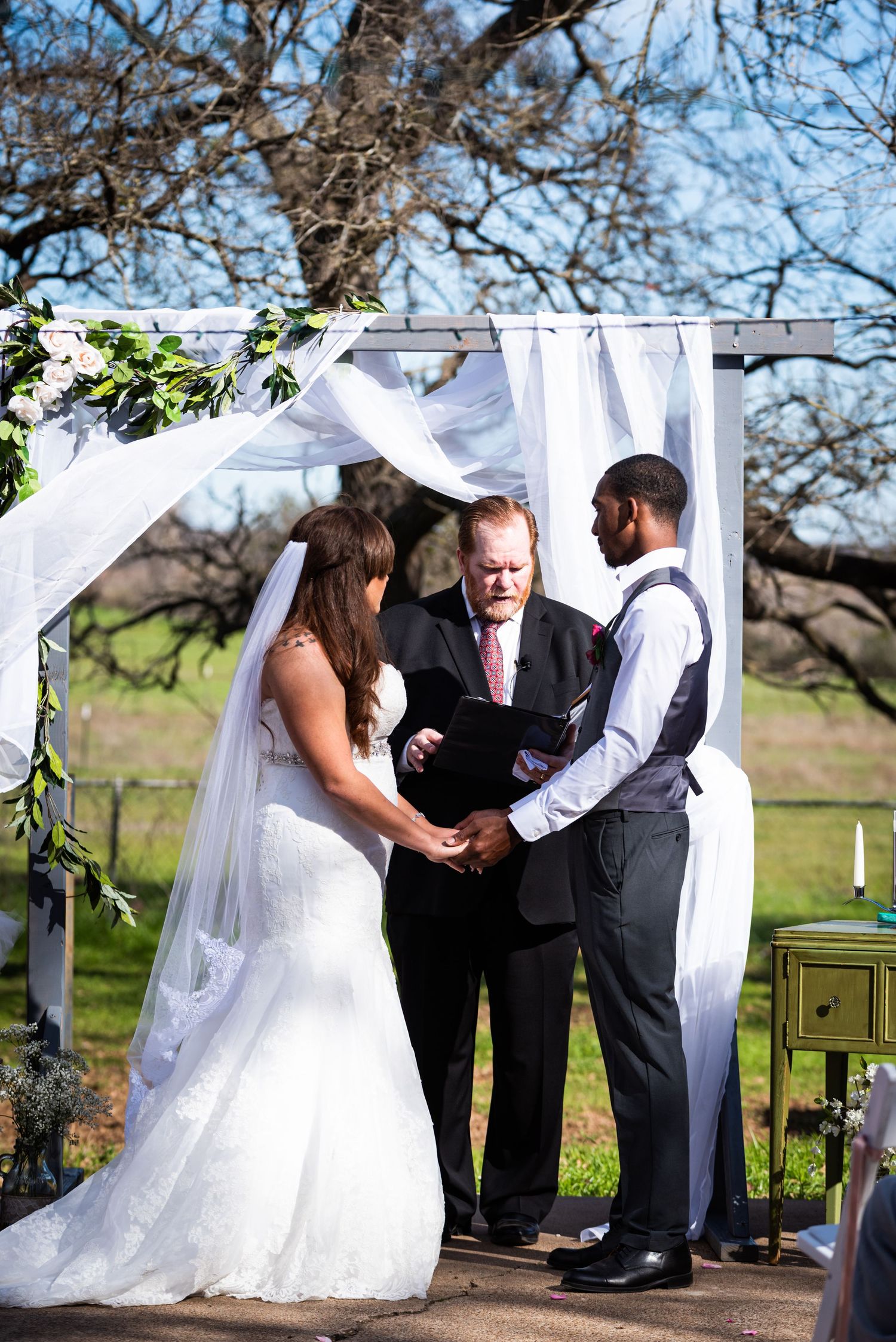 Outdoor wedding ceremony under a white arch with greenery and trees in the background.