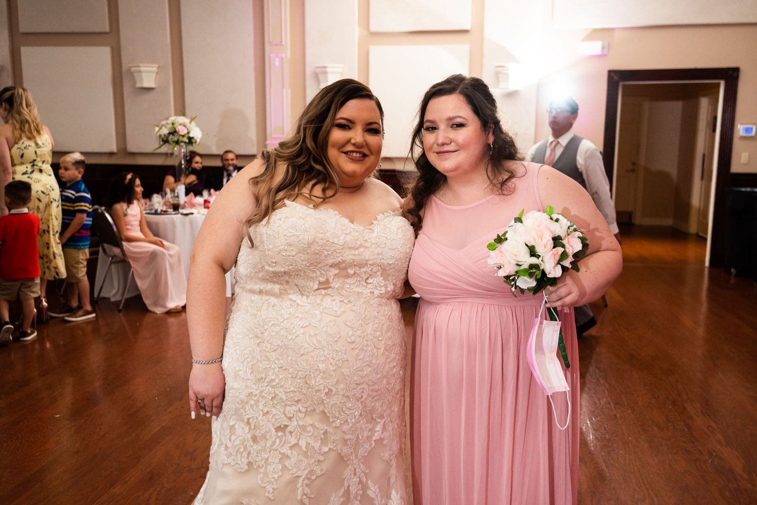 Two guests pose together at a formal event in a reception hall with hardwood floors.