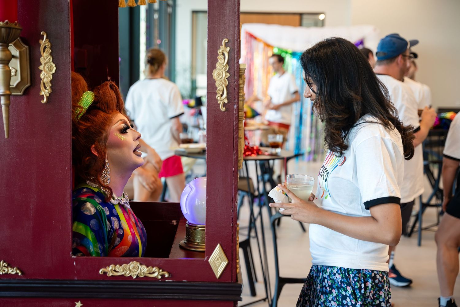 Drag performer in ornate booth interacts with a young woman at a colorful indoor Pride event.