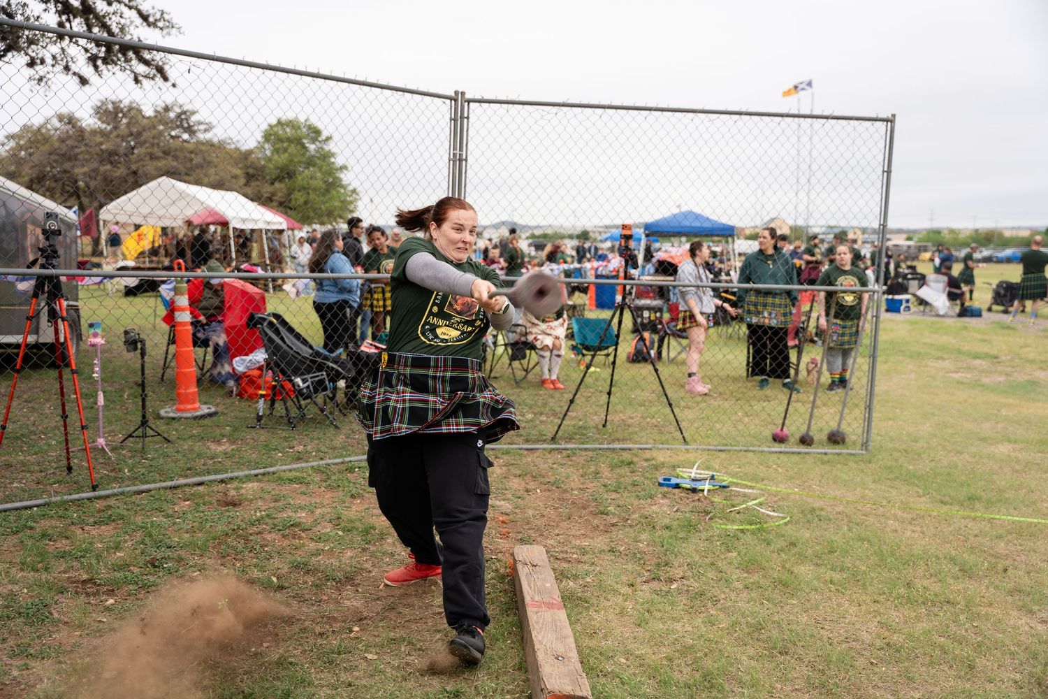 Woman in plaid vest drawing a compound bow at an outdoor archery competition with spectators and tents in background.
