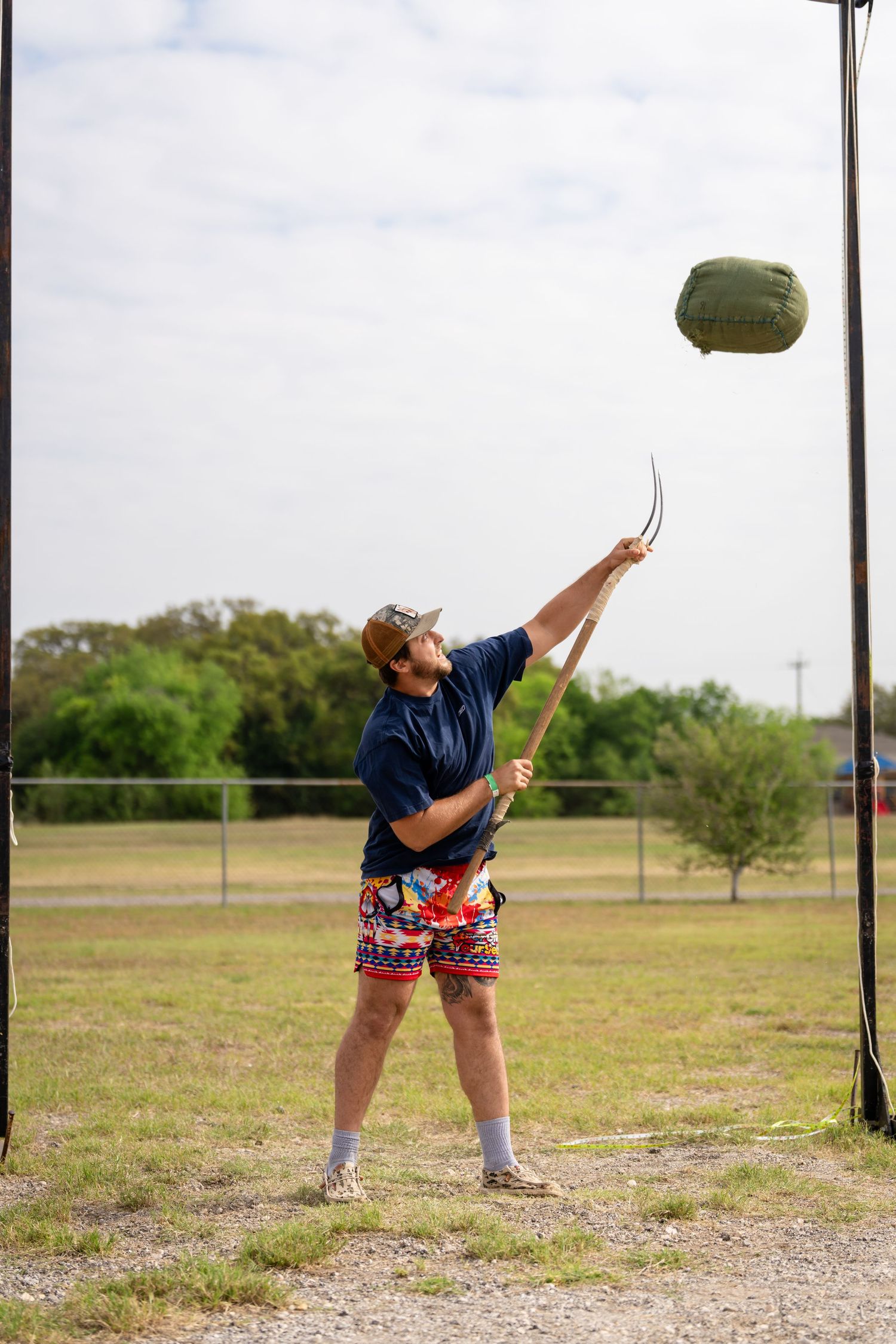 Man in colorful shorts and cap uses a pitchfork to toss a green bag in an outdoor field.