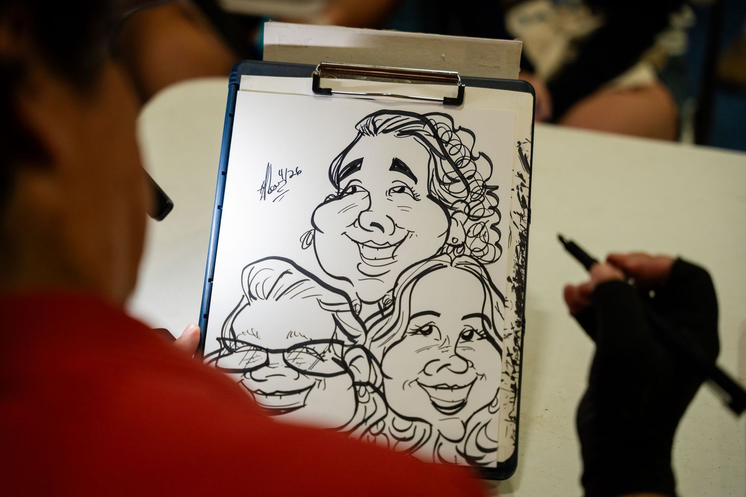 Artist drawing a caricature of two smiling women on a sketchpad at a live caricature event.