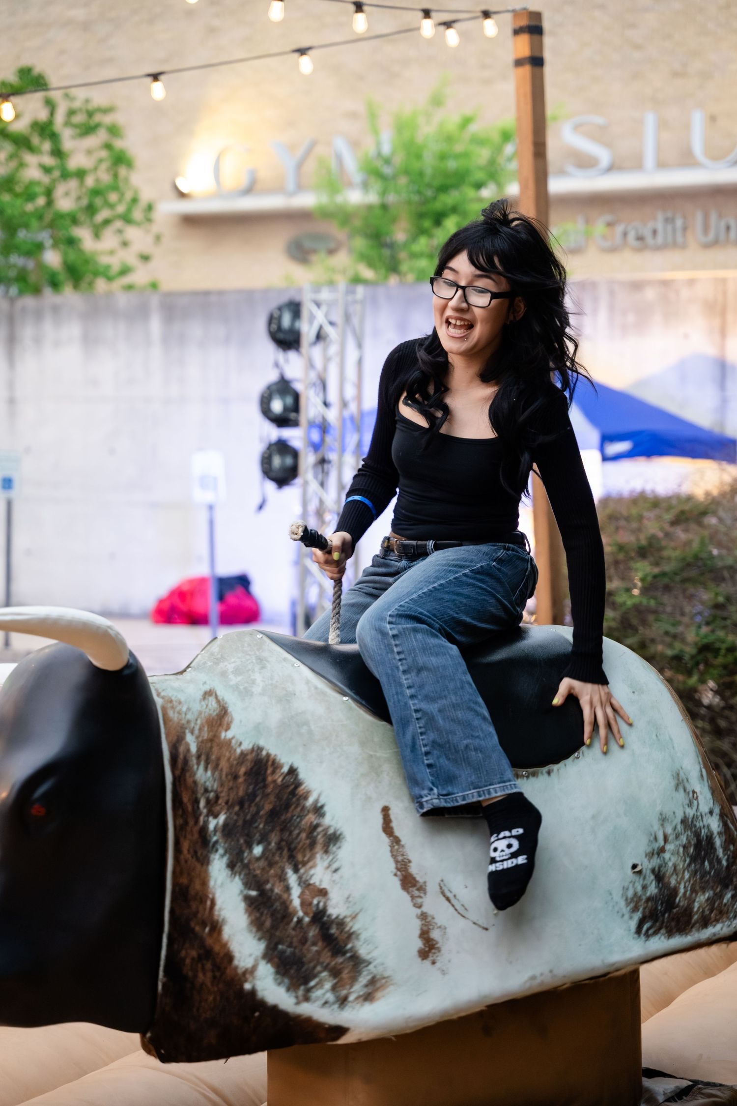 Woman riding a mechanical bull at an outdoor event, wearing jeans and black top, laughing with excitement.