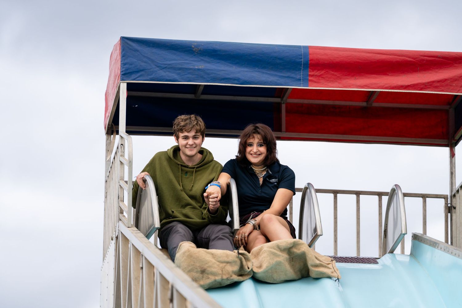 Two smiling people sit at the top of an outdoor fairground slide under a blue and red striped canopy.