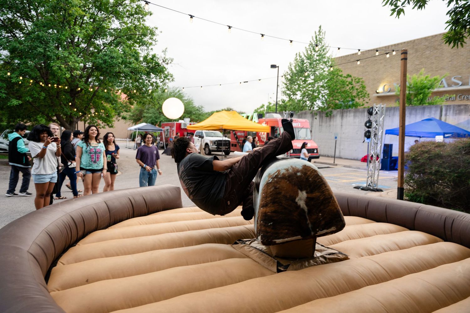 Person riding a mechanical bull at an outdoor festival event with spectators watching in the background.