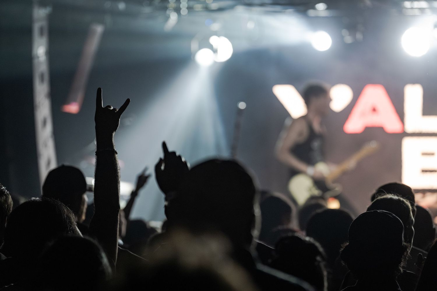 Crowd at a rock concert with devil horns hand gesture, guitarist performing on stage under bright lights.