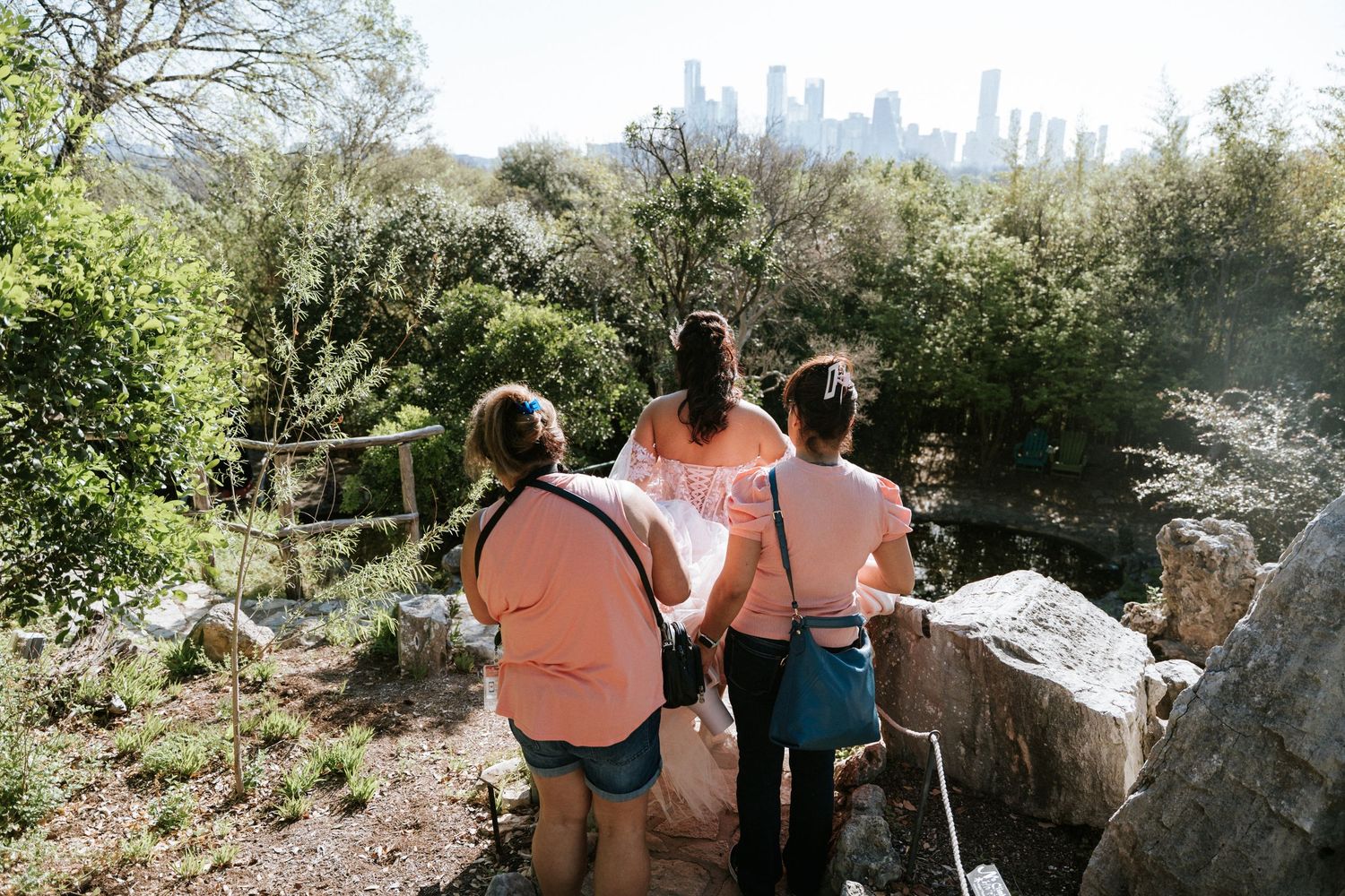 Three people stand on a rocky overlook surrounded by lush trees, gazing at a city skyline in the distance.