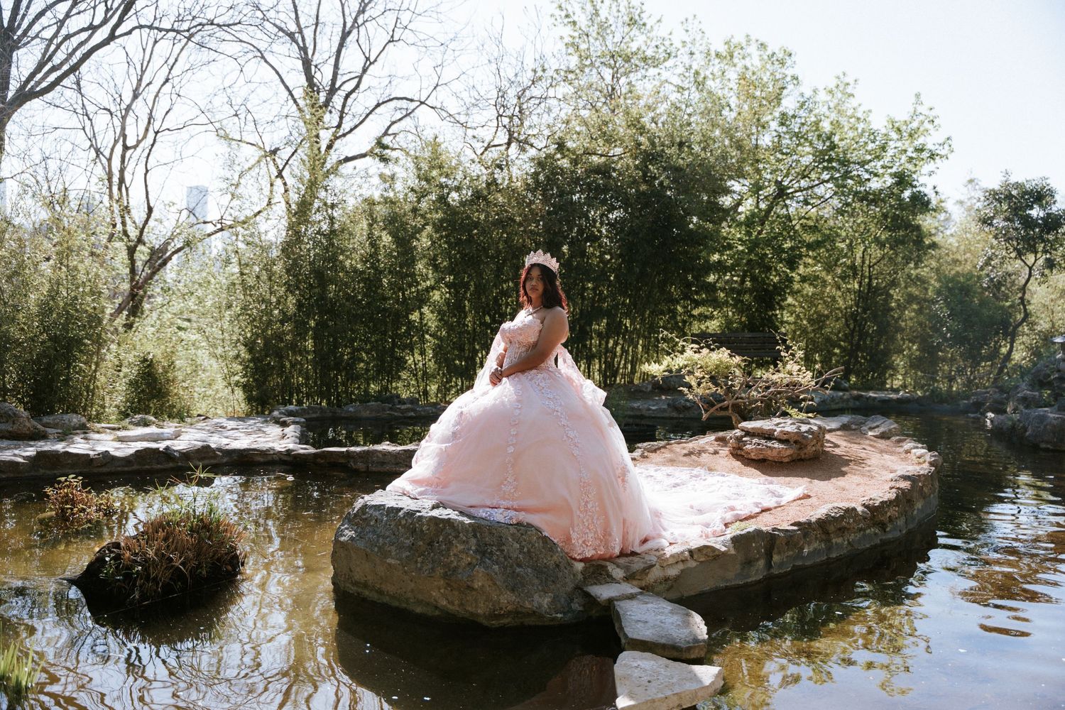 Young woman in a pink ball gown poses on rocks by a serene pond surrounded by lush trees.