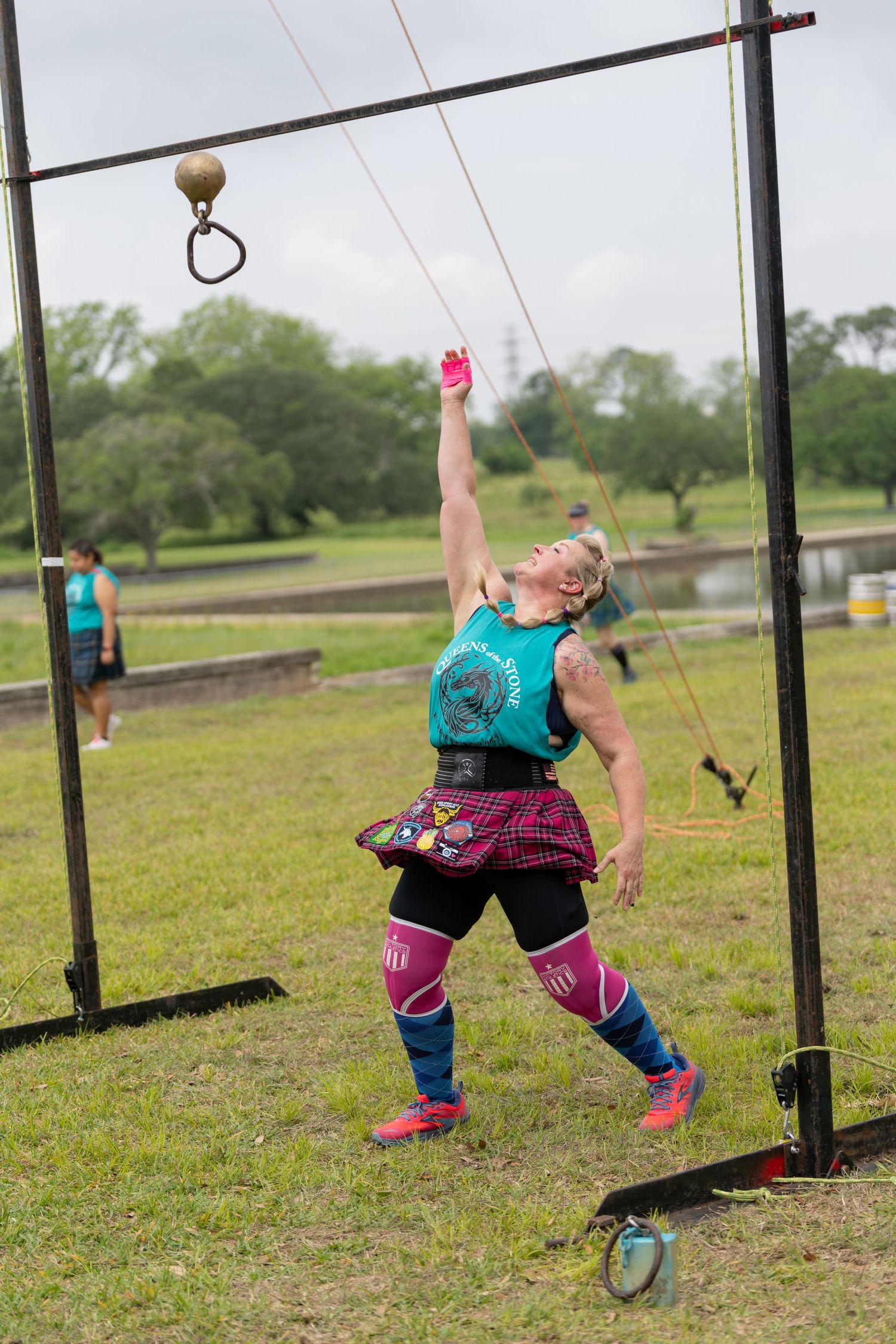 Woman in teal top and tartan skirt reaches up to ring a bell at an outdoor Highland Games competition.