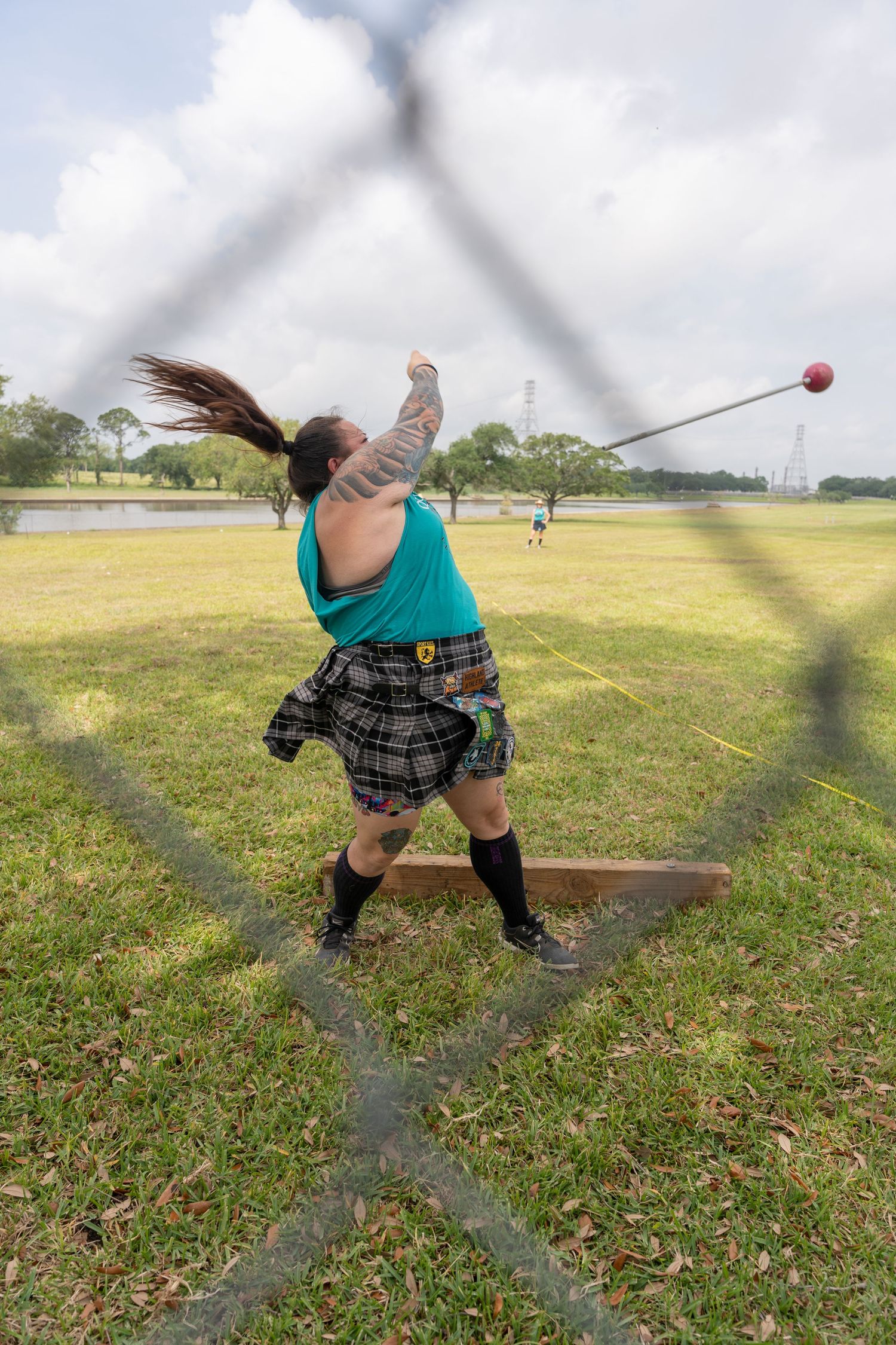 Woman in tartan kilt throws hammer at Highland Games competition, photographed through safety fence netting outdoors.