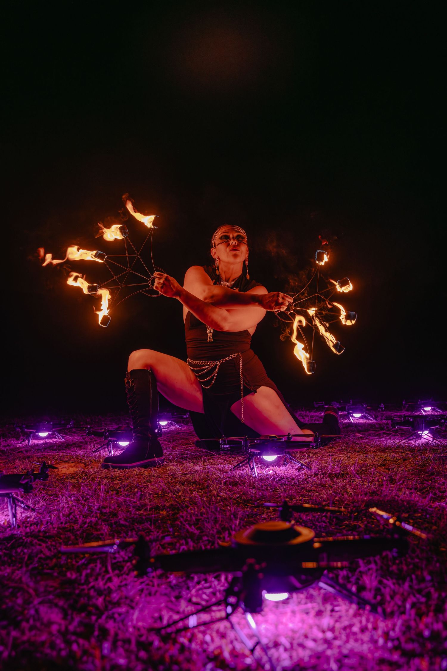Fire dancer performing at night with multiple flames, kneeling on grass surrounded by purple lights.