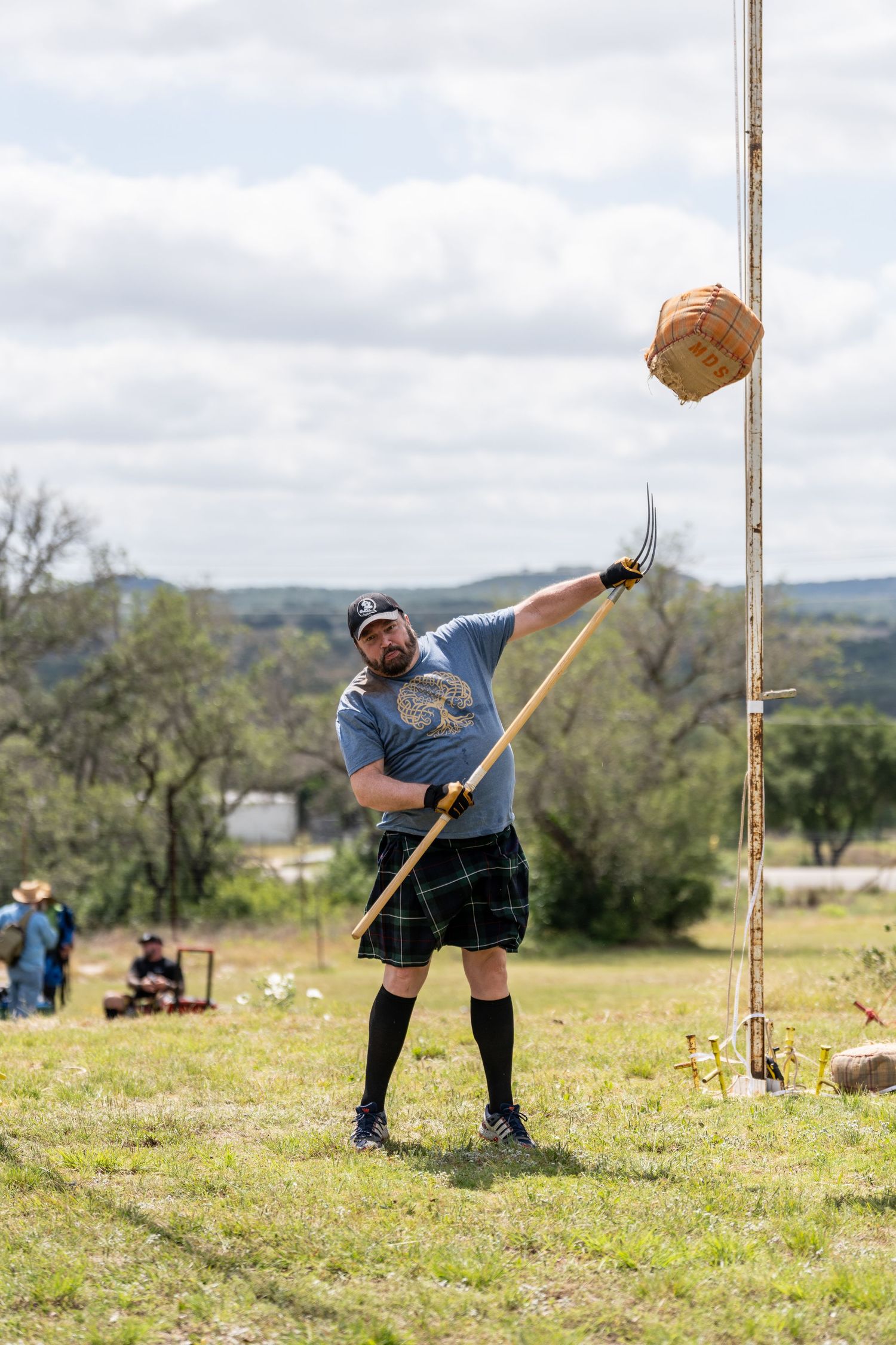 Man in kilt performs Scottish Highland Games sheaf toss with pitchfork at outdoor competition.