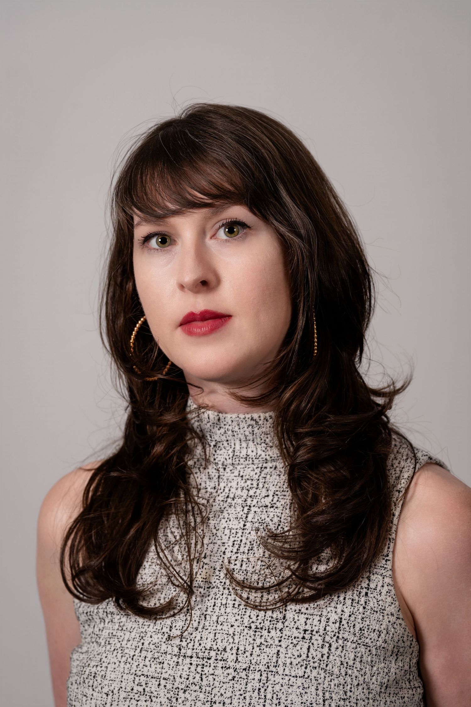 Young woman with dark hair, red lips, and gold hoop earrings wearing a sleeveless gray tweed top against a neutral background.