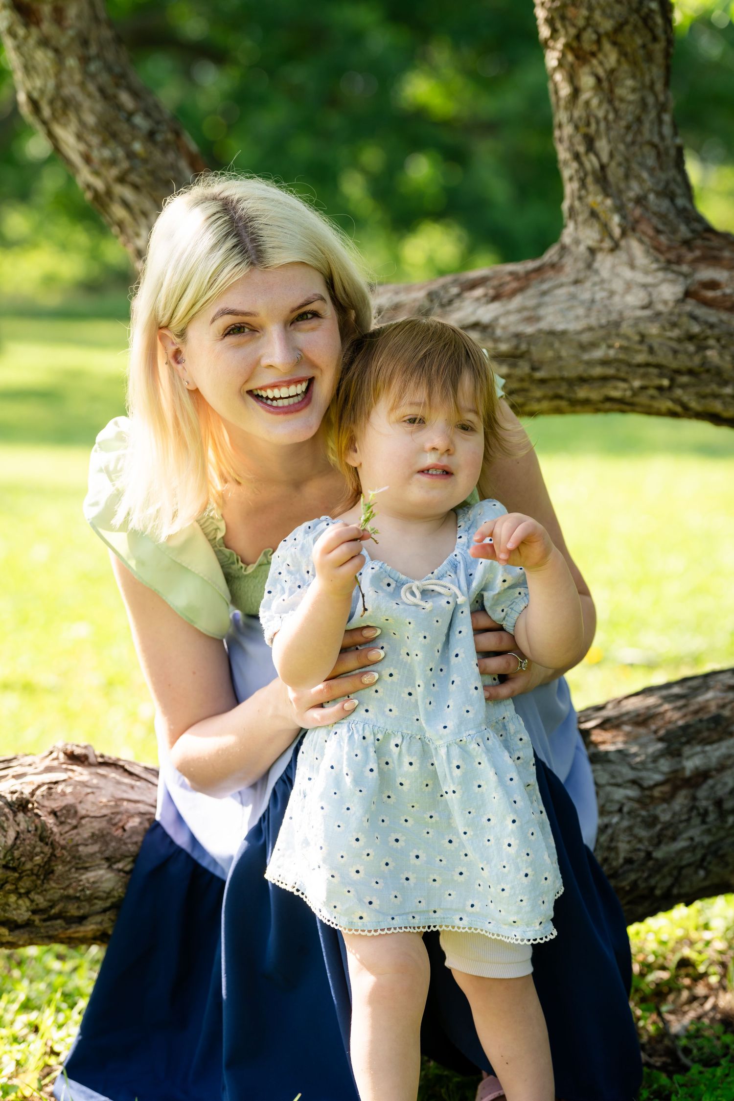 A laughing blonde mother hugs her toddler daughter in a sunny green park near a large tree.