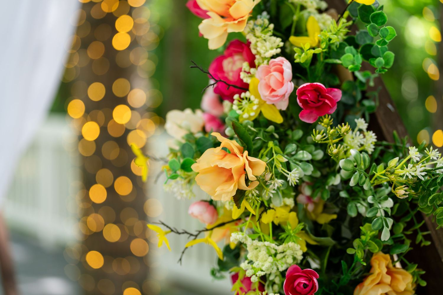 Colorful floral arrangement with pink, red, and yellow roses surrounded by lush green foliage with bokeh lights in background.