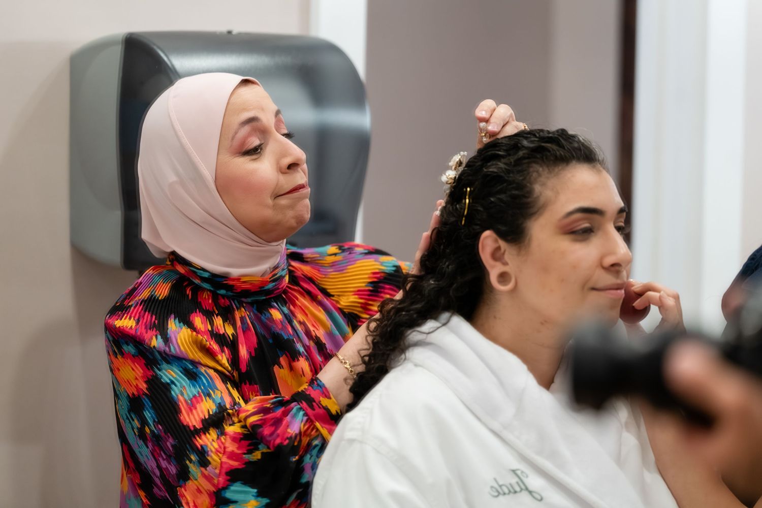 A hairstylist in a floral hijab styles a womans dark curly hair in a bright salon setting.