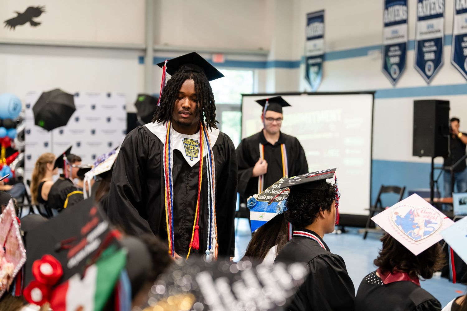 Graduate in black cap and gown with colorful honor cords walks among peers at graduation ceremony.