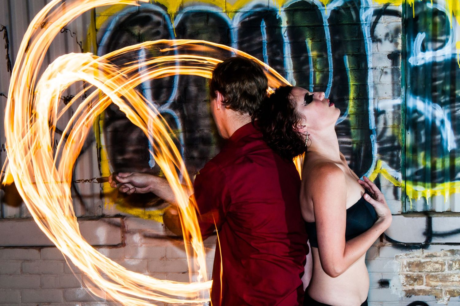 Fire performer creates glowing spiral trails of light against urban graffiti wall while person watches in background.