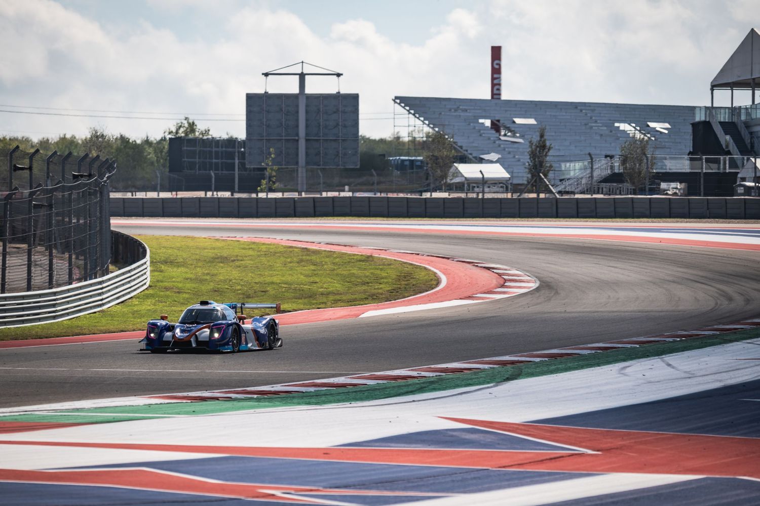 Race car speeds through a curving track with modern buildings and structures visible in the background.