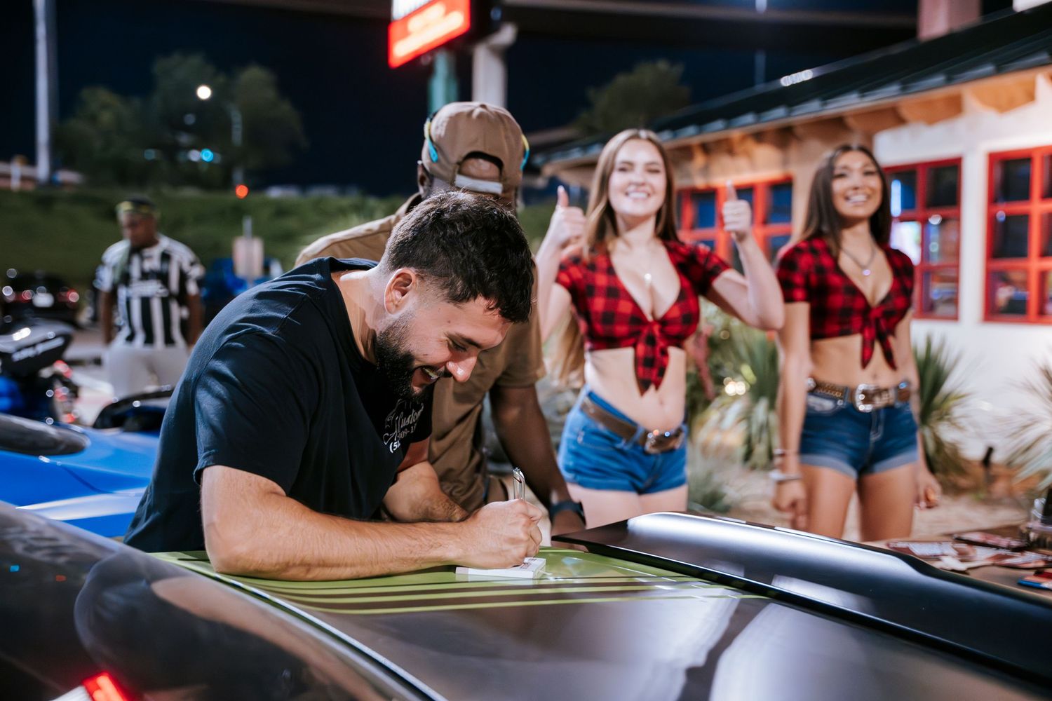 Person signs document on car hood while models in matching outfits watch at car meet.