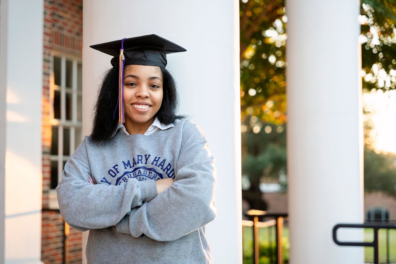 Graduate in academic regalia stands between white columns in university sweatshirt.