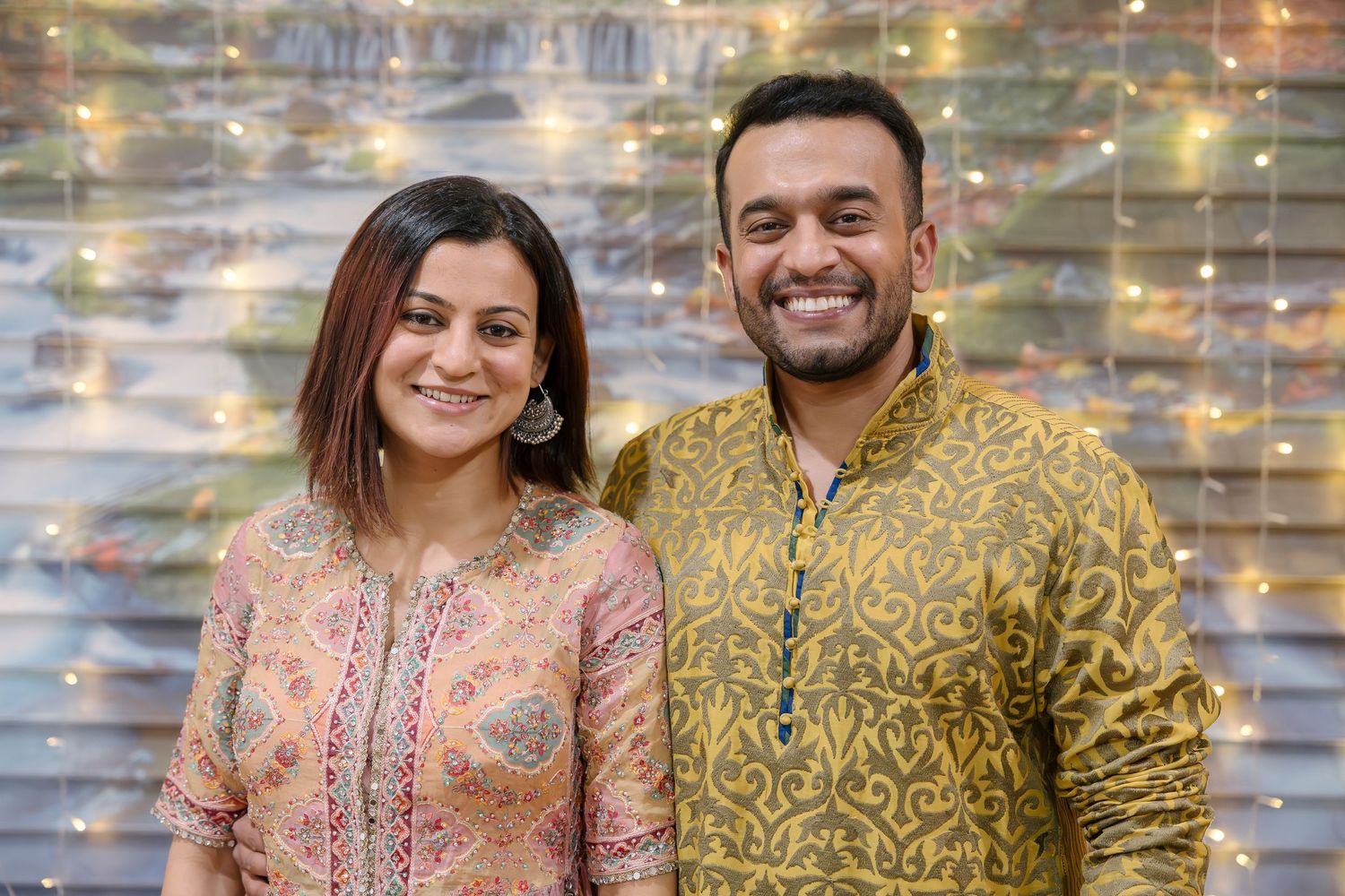 A couple in festive Indian attire posing against a shimmering backdrop.