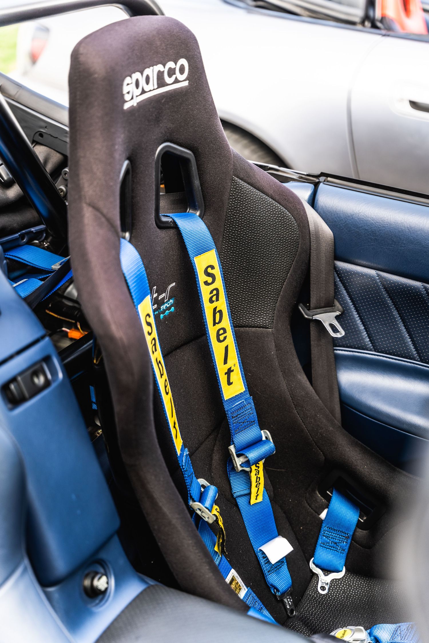Black Sparco racing seat with blue safety harness belts installed in a sports car interior.