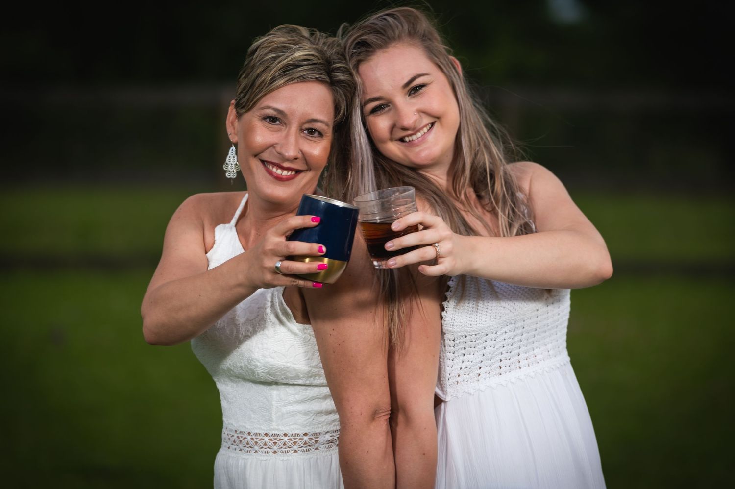 Two women in white dresses smile while holding drinks at an outdoor celebration.