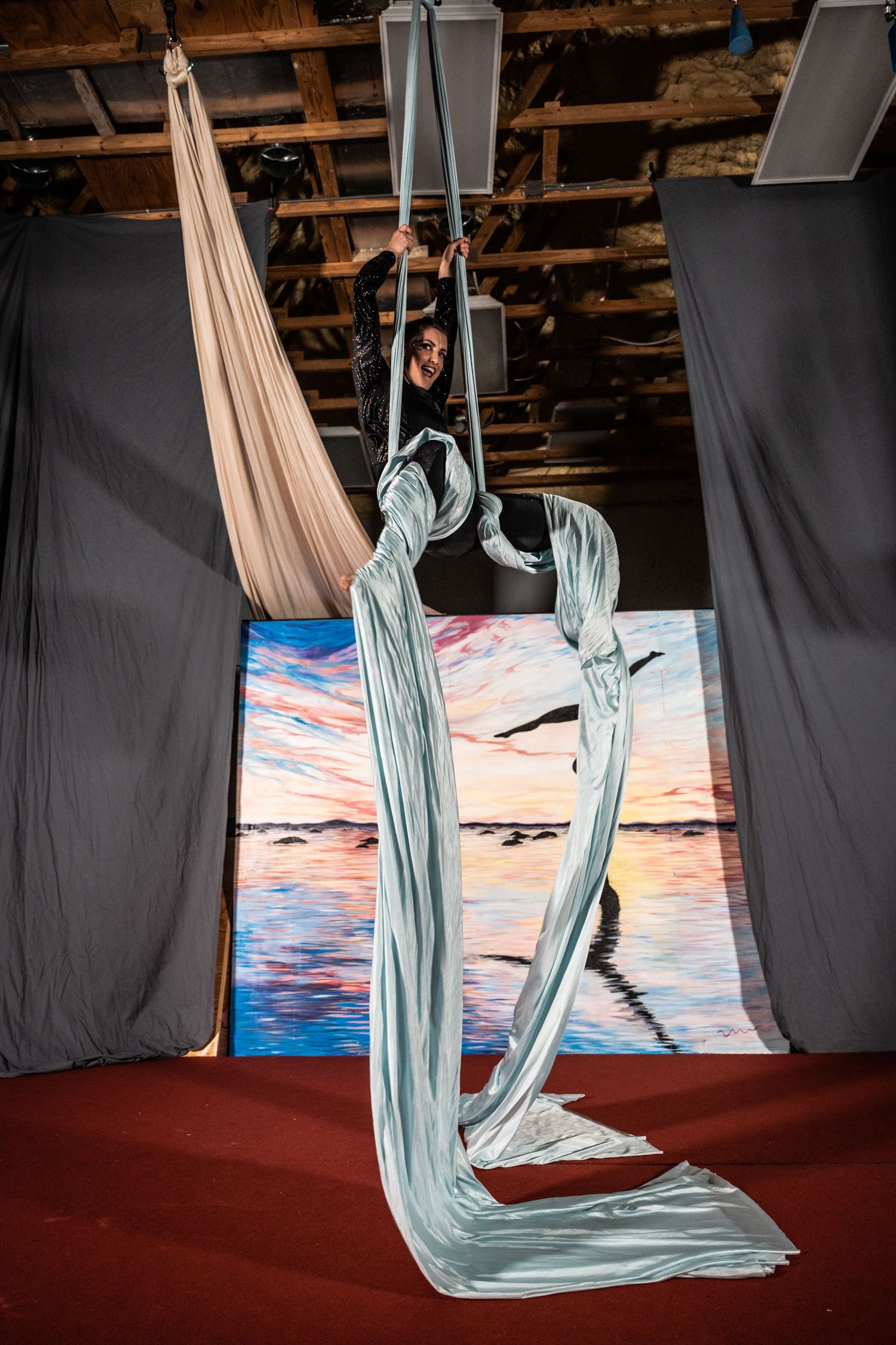 Aerial silk artist performs graceful moves with light blue fabric against sunset backdrop.