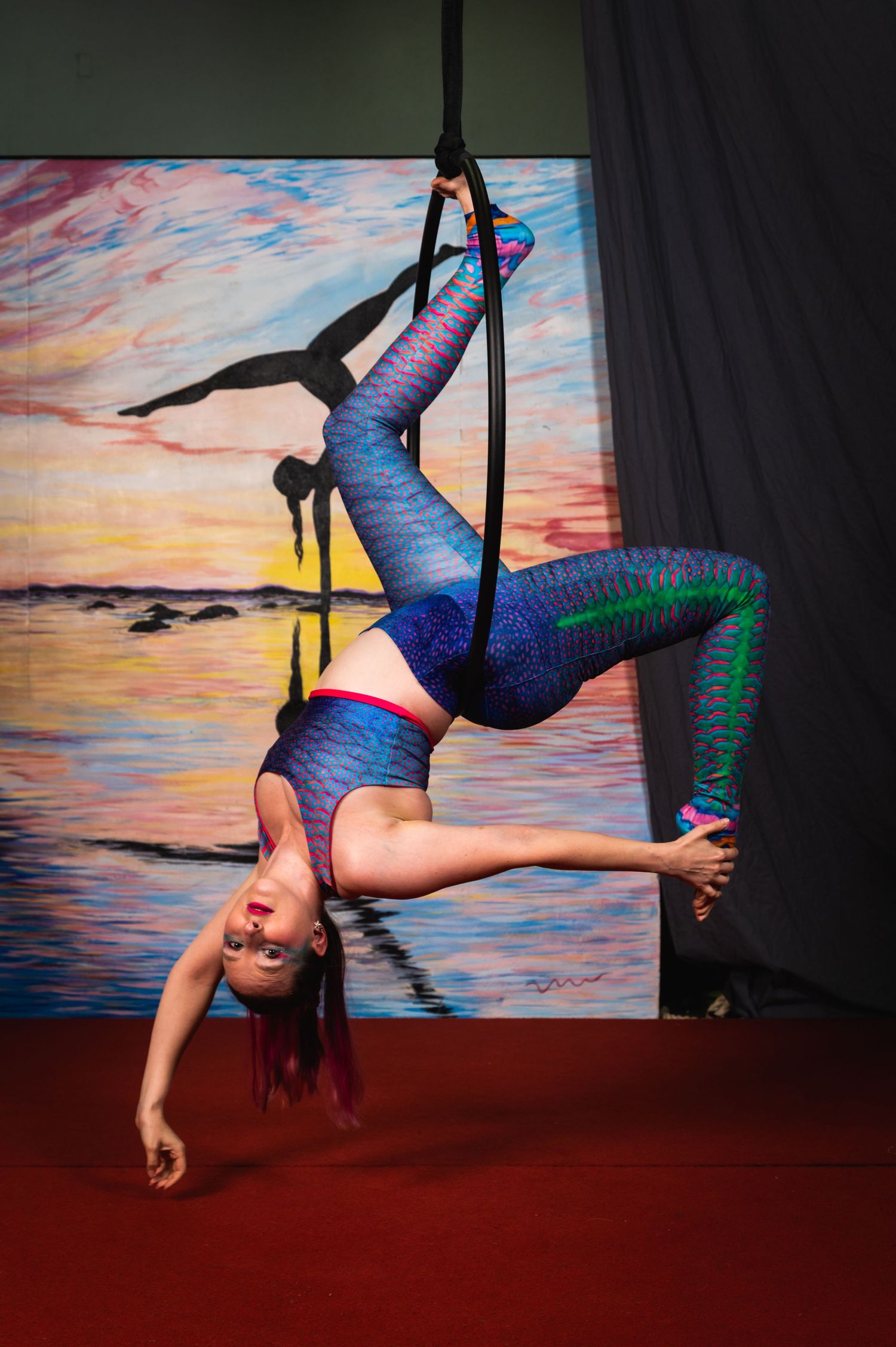 Aerial performer in blue and green sparkly outfit hangs upside down on aerial hoop against sunset backdrop.