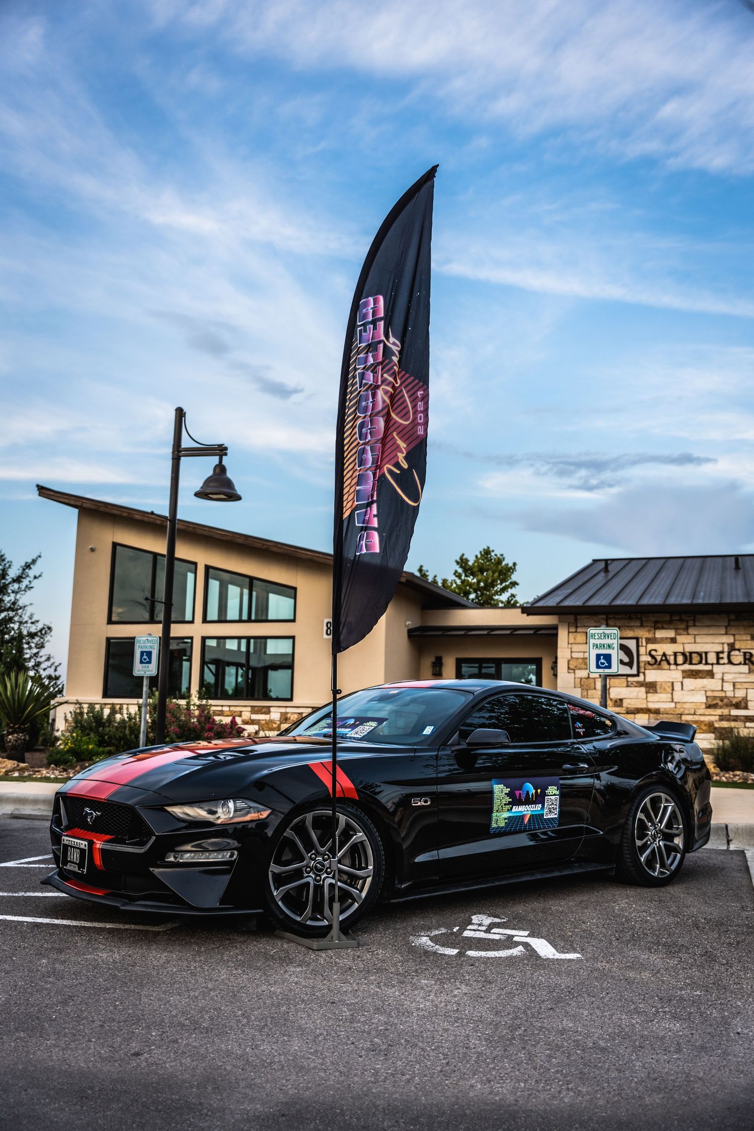 Black Ford Mustang with red racing stripes parked near a promotional feather flag banner.