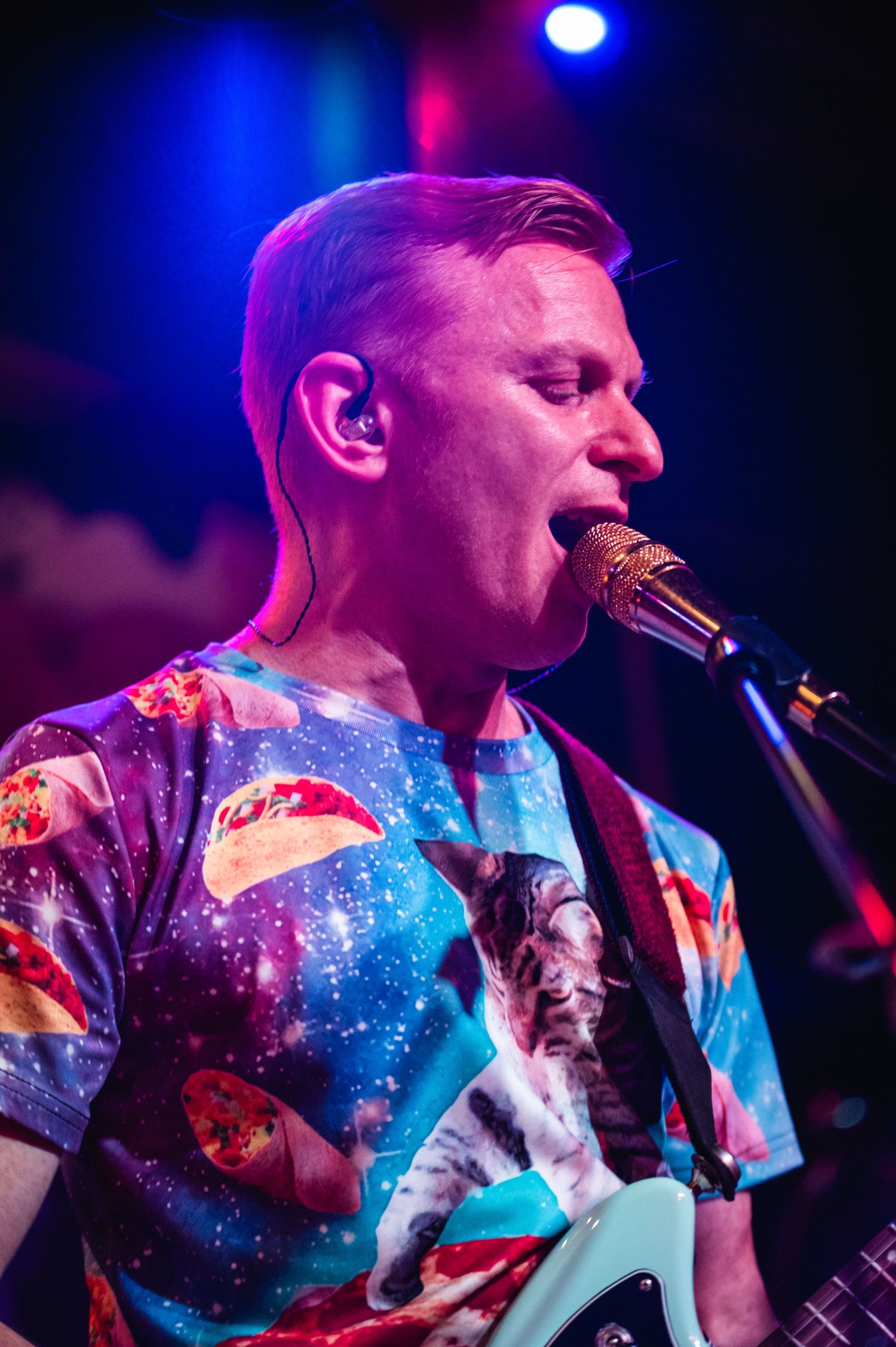Close-up of performer singing into microphone wearing cosmic-themed t-shirt under blue and red lights.