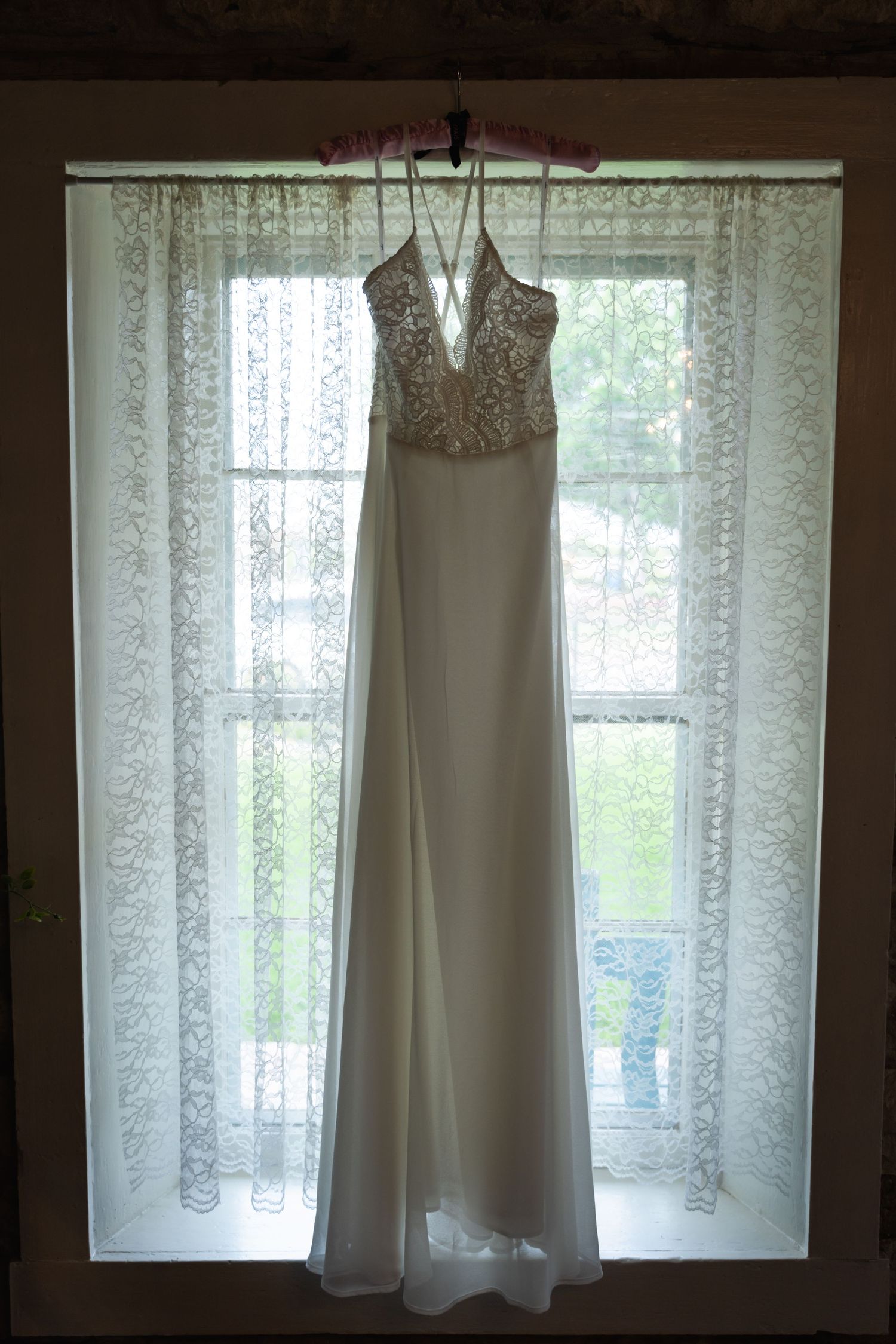 White wedding dress hanging in window with lace curtains creating silhouette effect.