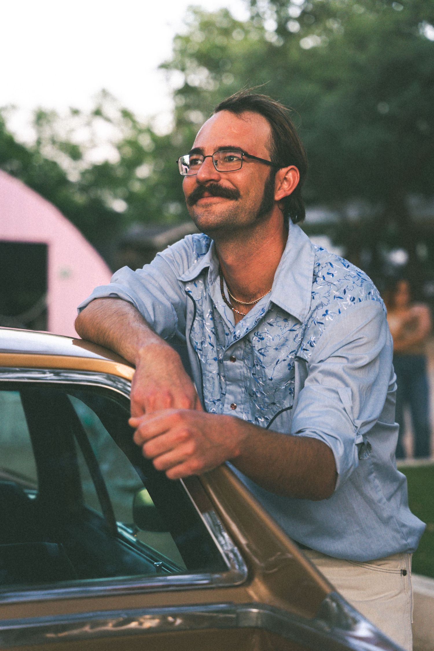 Casual portrait of someone leaning on a vintage car at sunset wearing glasses and a denim shirt.