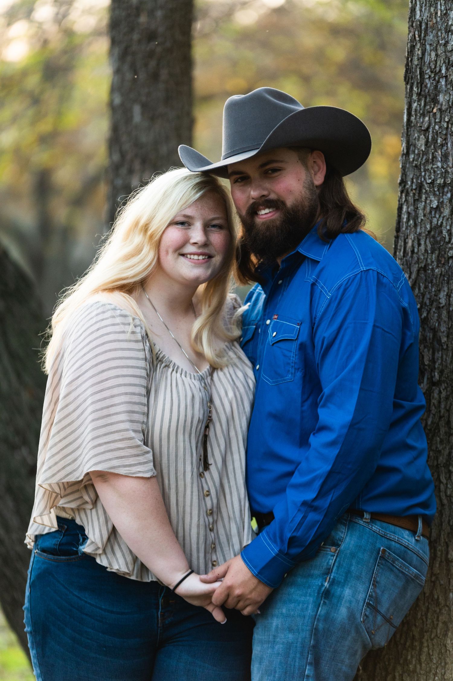 A couple in casual western attire poses against a tree trunk during an outdoor photo shoot.