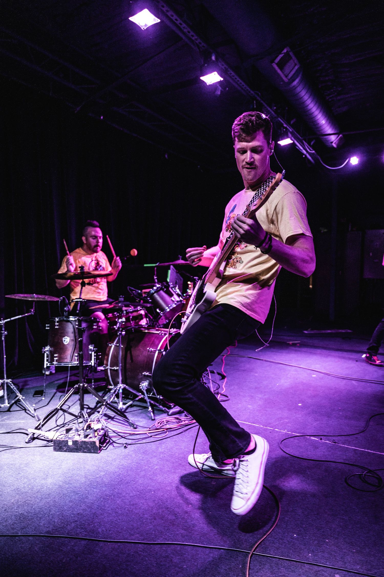 Musicians perform on stage with purple lighting and drum kit at a live concert venue.