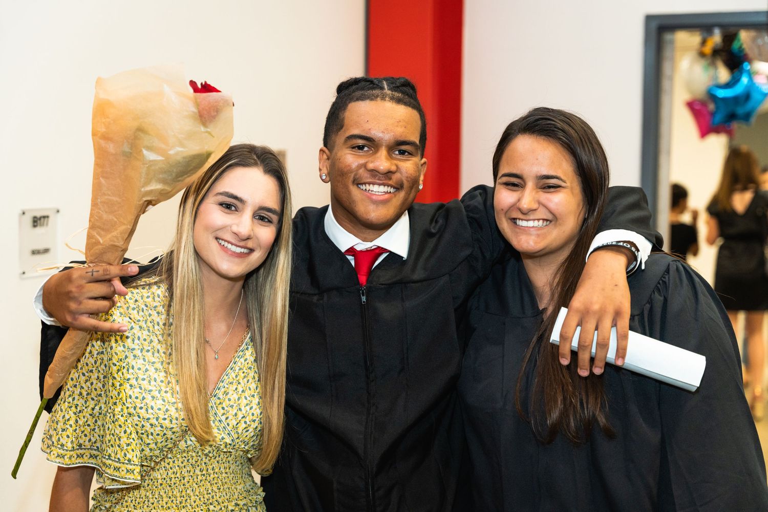 Three graduates celebrate together at commencement ceremony with flowers and diplomas.