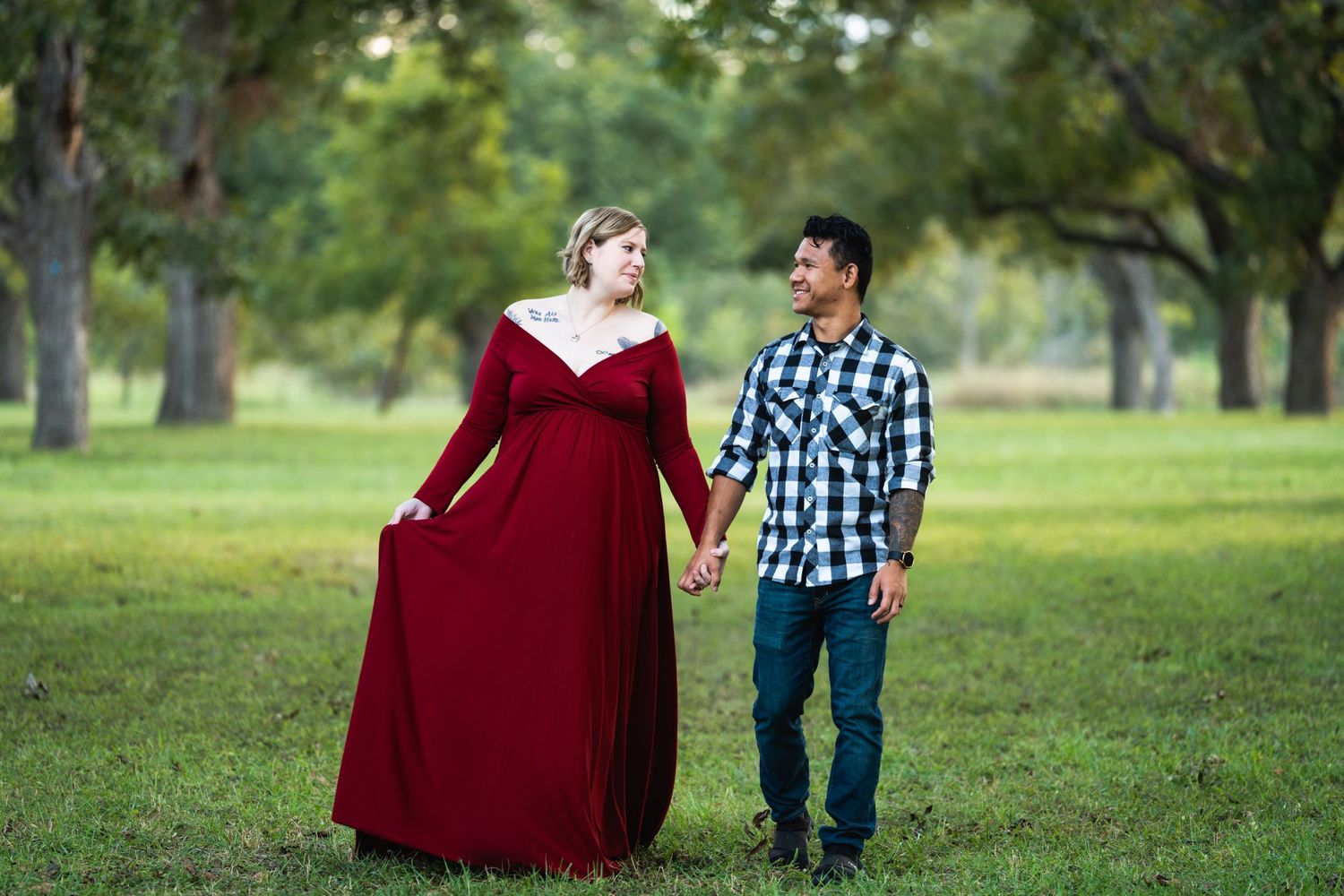 Couple standing together outdoors, woman wearing a long red dress in a park setting.