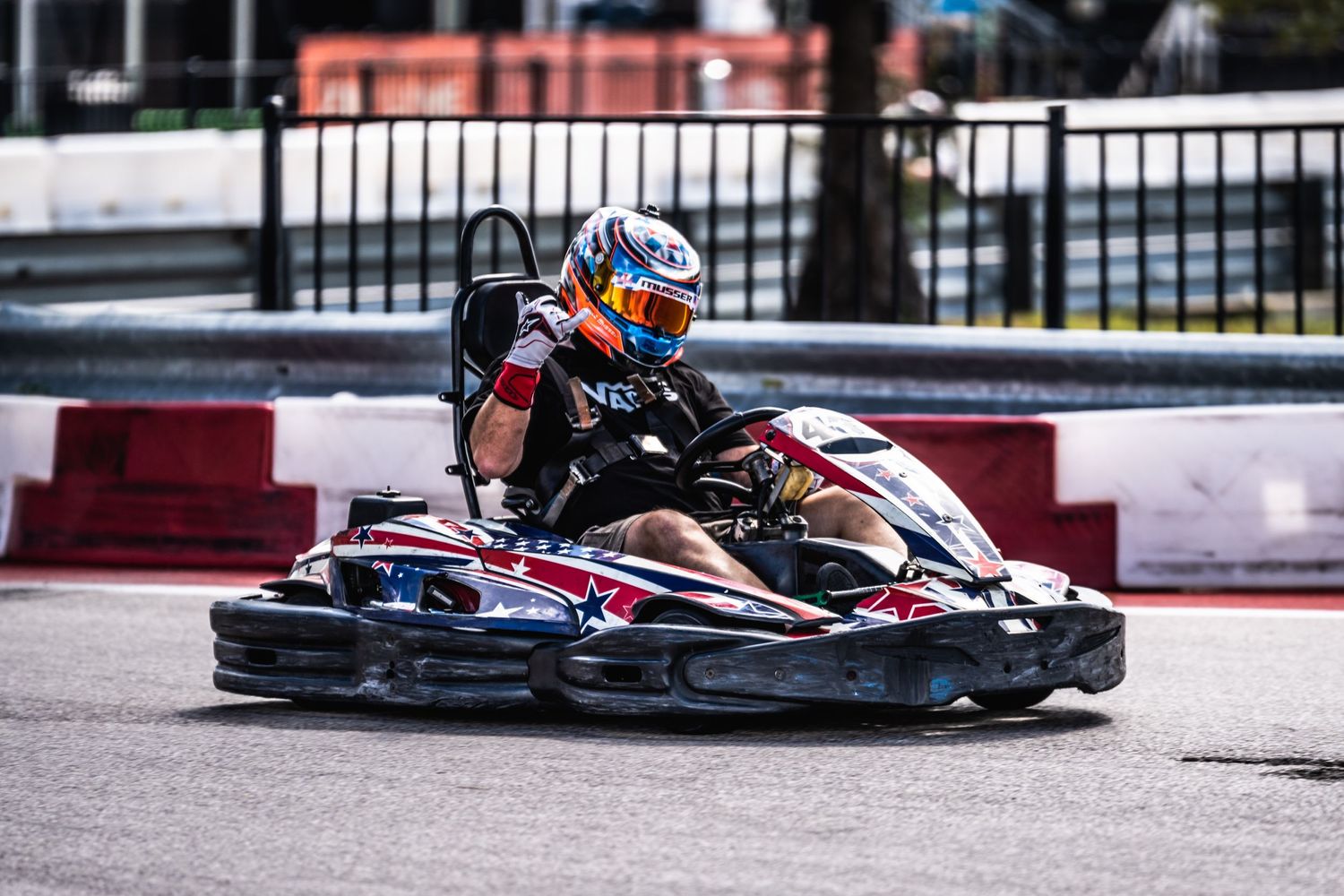 Go-kart racer taking tight corner on professional racing circuit with red and white barriers.